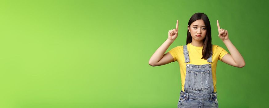 Pity cute sad asian girl brunette, grimacing pulling sorrow upset face frowning bothered, pointing fingers up regret awful situation, indicate top promo, stand green background unhappy.
