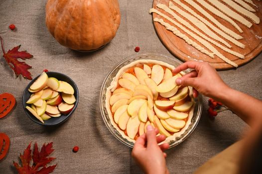 Top view of housewife, chef confectioner putting slices of apples on a raw pumpkin pie, preparing homemade traditional American classic tarte with crust lattice, for Thanksgiving Day. Autumn holidays