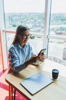 Happy freelancer girl in classic glasses looking at phone and smiling while sitting in modern coworking space, carefree millennial woman in glasses enjoying leisure time for communication