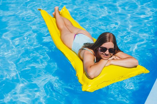 Summer lifestyle portrait of beautiful woman in sunglasses floating on inflatable mattress in outdoor swimming pool. Summer vacation on a tropical island.