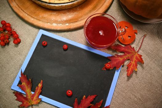 Top view of a blank chalk board with copy advertising space for a promotional text, ripe red viburnum berries, red maple autumn leaves and a glass cup of hot mulled wine on the linen tableclot