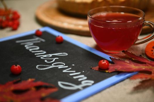 Selective focus. Still life with a glass teacup with healthy antioxidant hot herbal drink, blurred foreground with dry fallen autumn maple leaves and a blackboard with lettering Thanksgiving Day.