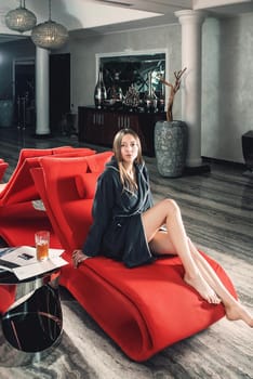 Portrait of young beautiful woman with long sexy legs relaxing in a fashionable red chair in a gray bathrobe, waiting for treatment. Luxery spa center