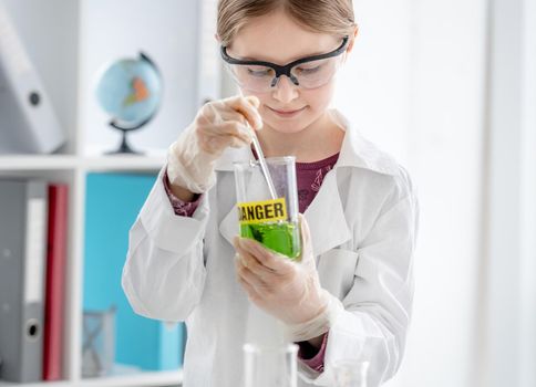 Smart girl doing scientific chemistry experiment wearing protection glasses, and add liquid to bottle with title danger. Schoolgirl with chemical equipment on school lesson