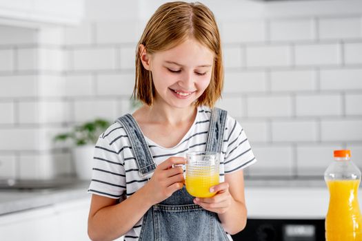 Preteen girl wih orange juice in glass smiling at kitchen. Pretty child kid with vitamin fruit drink happy at home