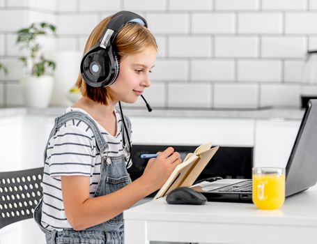 Preteen girl wearing headphones study with laptop during online e-learning lesson at home at pandemic coronavirus. Child pupil making school video call with orange juice glass on table