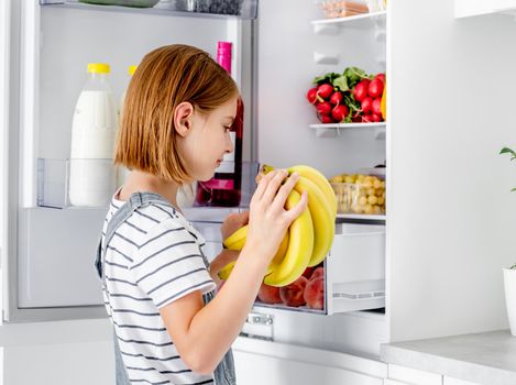 Preteen girl with bananas looking at fridge with vitamin healthy food at kitchen. Pretty child kid holds yellow fruits at home