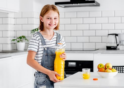 Preteen girl with orange juice bottle and glass at kitchen. Pretty child kid with vitamin fruit drink at home