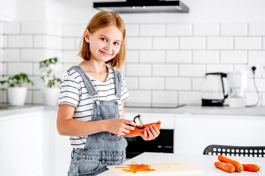 Preteen girl pilling carrot at kitchen and smiling. Child with orange vegetables food cooking at home