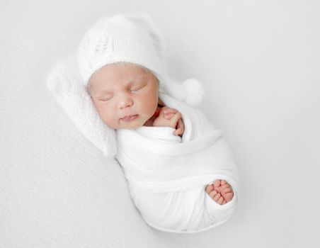 Newborn baby girl swaddled in white fabric and wearing hat sleeping. Infant child kid studio portrait