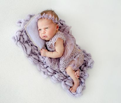 Newborn baby girl wearing wreath and knitted costume lying on pillow and looking back. Infant child kid studio portrait