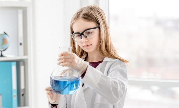 Smart girl doing scientific chemistry experiment wearing protection glasses, holding bottle and measuring blue liquid. Schoolgirl with chemical equipment on school lesson