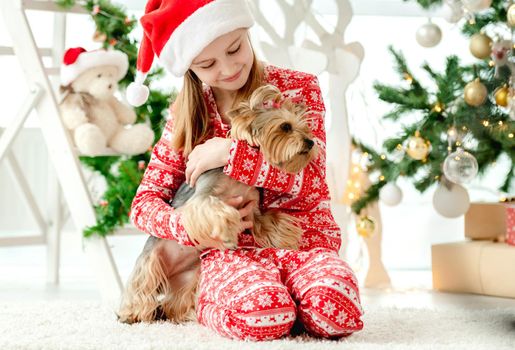 Child girl hugging dog near Christmas tree and happy smiling. Kid wearing Santa hat celebrating New Year with doggy pet terrier