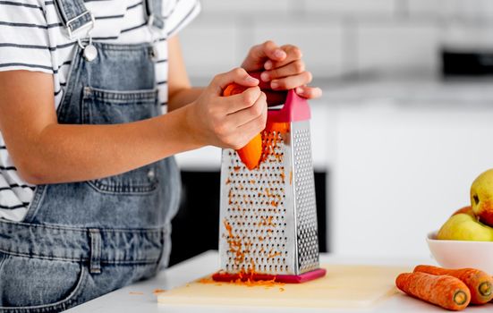 Girl child hands grinds carrot using grater at kitchen closeup. Pretty kid cooking vegetables at home