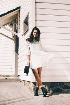 Sunny lifestyle fashion portrait of young stylish hipster woman walking on the street, wearing trendy white dress, black hat and boots. White wooden house on a backgrond.