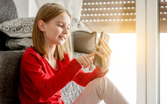 Child girl holds smartphone in her hand and smiles chatting with friends. Cheerful kid spends time with cellphone sitting on floor near sofa by window.