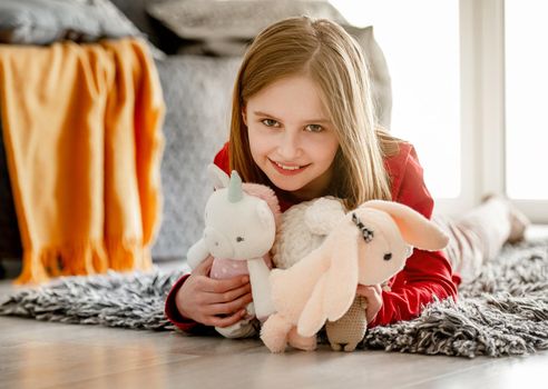 Happy little girl holds toys and lies on soft carpet smiling. Cheerful child spends time on floor at home