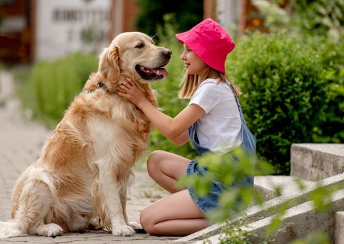 Preteen girl wearing hat hugging golden retriever dog sitting outdoors in summertime. Pretty kid petting fluffy doggy pet in city