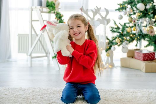 Child girl in Christmas time sitting on carpet in room with decorated tree and gifts. Pretty kid at home in New Year holidays