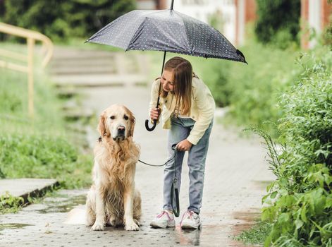 Preteen girl with golden retriever dog in rainy day walking outdoors in city park under umbrella. Cute kid child with labrador pet doggy outside