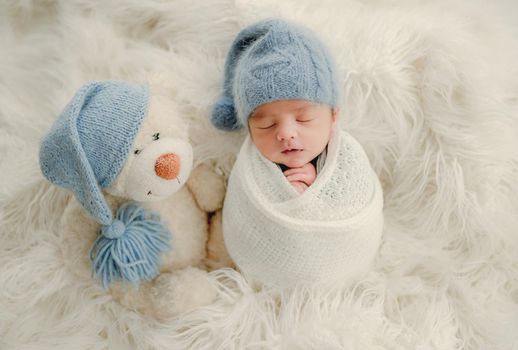 Newborn baby boy with knitted snowman toy sleeping swaddled in fabric on white fur. Infant child kid Christmas and New Year holiday portrait