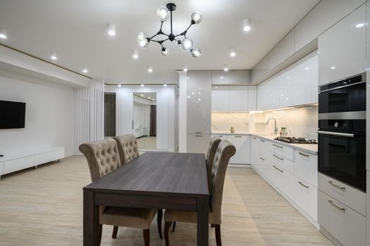 Interior of a huge white luxury kitchen in a studio apartment with tv and oven, lots of copy space, closeup view to dining table with chairs