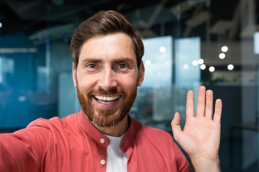 Young happy businessman looking into smartphone camera and talking on video call, man working inside modern office building, smiling and talking remotely with friends, using communication app.