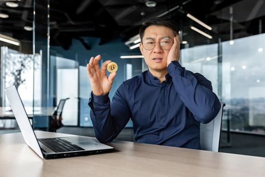 Dissatisfied and confused in depression asian financier investor, holding coin money crypto currency bitcoin in hands, male businessman working dissatisfied with exchange rate, broker in despair.