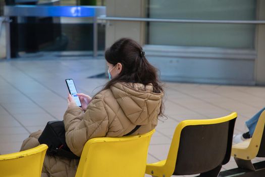 a young woman in a warm down jacket with green screen smartphone sits on an armchair in the waiting room in the subway watches the train and use social media, . High quality photo