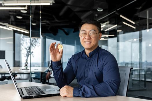 Portrait of satisfied asian broker investor, businessman looking at camera and smiling man holding crypto currency bitcoin virtual money in hands, working inside modern office building.