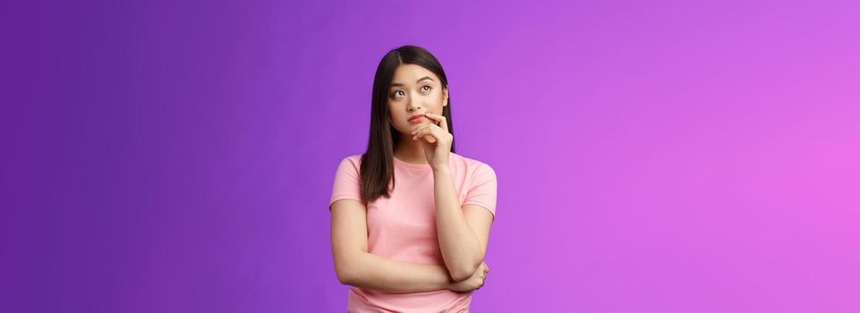 Thoughtful creative stylish asian girlfriend thinking-up perfect b-day gift, touch chin thoughtful look upper left corner, pondering, imaging in mind ideas, stand purple background.