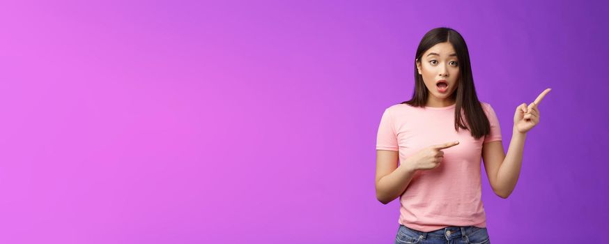 Excited alarmed cute asian female describe shocking scene pointing right, show index fingers sideways thrilling event, discuss amazing performance look camera worried, stand purple background.