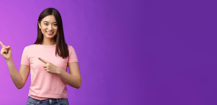 Friendly cute pleasant asian woman showing way, indicating interesting promo, pointing left smiling broadly, introduce advertisement product, stand purple background cheerful. Copy space