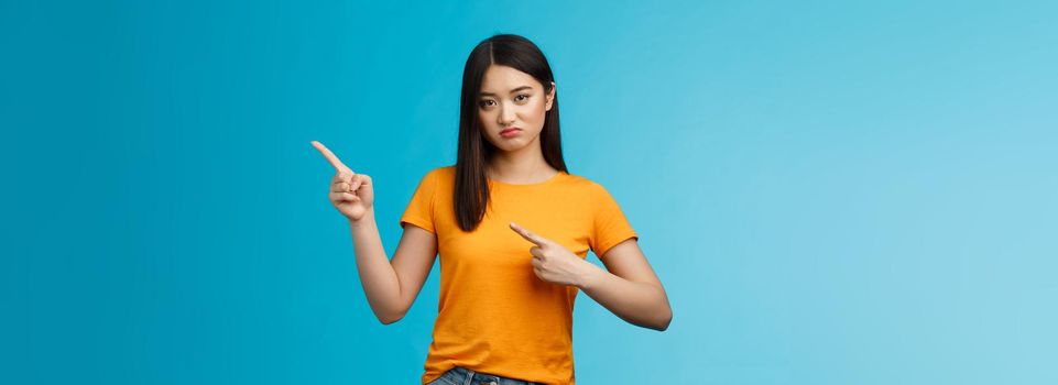 Picky disappointed asian girl with unsatisfied face, pointing upper right corner, sulking offended, feel jealous friend went summer vacation abroad alone, stand blue background displeased.