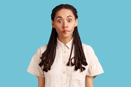 Surprised young woman with dreadlocks makes fish lips, being not scared to show emotions, tries to make her crying friend laugh, wearing white shirt. Indoor studio shot isolated on blue background.