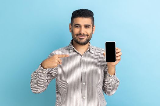 Cheerful beaded businessman pointing finger at smartphone with empty screen, looking at camera with smile, freespace for adv, wearing striped shirt. Indoor studio shot isolated on blue background.