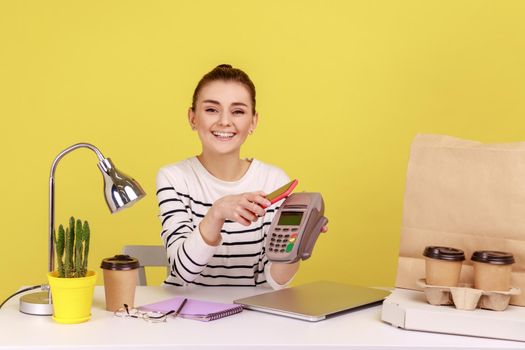 Happy young woman sitting at workplace with mobile smart phone and paying by pay pass pos. contactless, nfc technology on cellphone and payment. Indoor studio studio shot isolated on yellow background