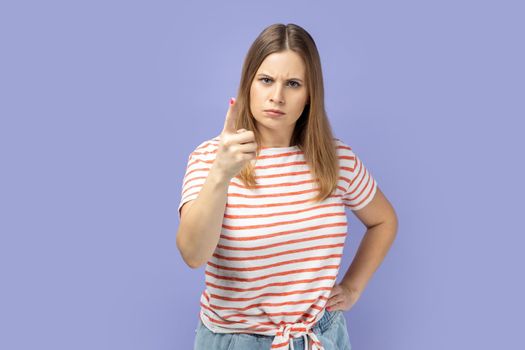 Portrait of blond woman wearing striped T-shirt pointing finger at camera and looking with dissatisfied suspicious expression, warning about troubles. Indoor studio shot isolated on purple background.