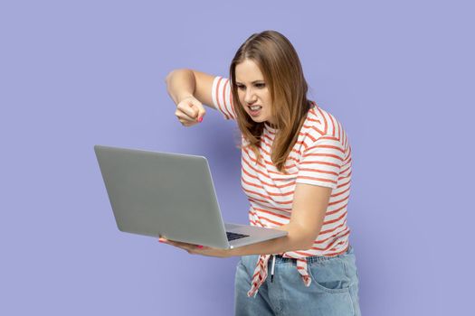 Portrait of angry aggressive blond woman wearing striped T-shirt working on laptop, having online conversation, boxing to notebook display. Indoor studio shot isolated on purple background.