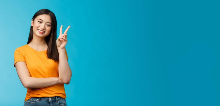 Optimistic cute tender asian young millennial girl send positive vibes, tilt head show peace, victory sign, smiling broadly, wish fantastic holidays, stand blue background joyful upbeat.