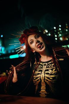 Female with skeleton makeup at a Halloween party make grimaces and looks at camera. Celebration in a nightclub. Woman in creepy costume of Mexican goddess of death. All Saints' Night.