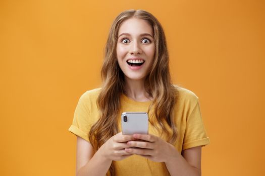 Lifestyle. Waist-up shot of amused and excited cute young woman with wavy hairstyle smiling broadly astonished and amazed reacting to awesome smartphone holding in hands posing delighted over orange wall.