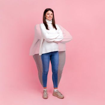 Full length portrait of happy satisfied woman wearing jumper and jeans standing with folded arms, showing herself before and after losing weight. Indoor studio shot isolated on pink background.