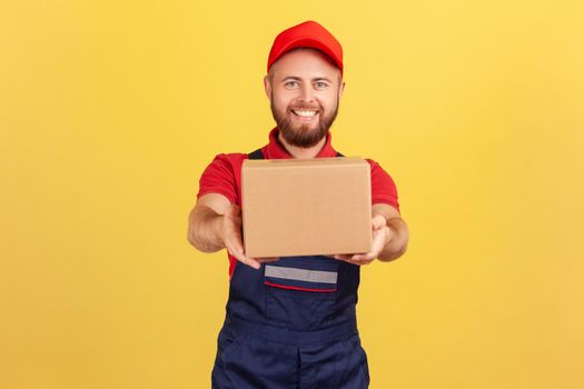 Portrait of smiling courier man holding cardboard parcel, delivering order door-to-door, shipment and cargo transportation service. Indoor studio shot isolated on yellow background.