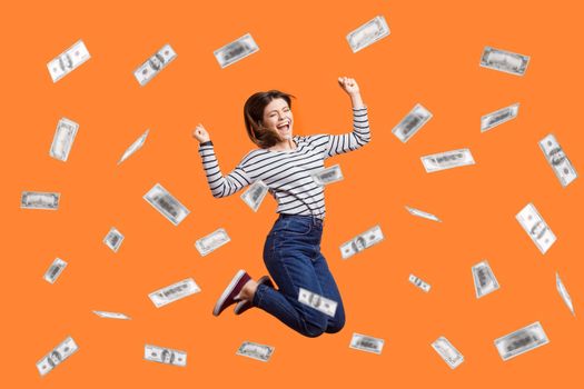 Portrait of satisfied excited woman jumping with clenched fist, celebrating winning lottery, flying in money rain, dollars falling. Indoor studio shot isolated on orange background.