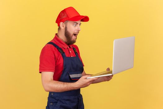 Side view portrait of shocked astonished handyman standing laptop in hands and working online, getting huge order, wearing uniform and red cap. Indoor studio shot isolated on yellow background.