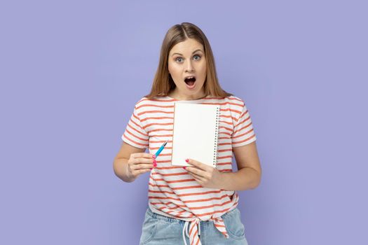 Portrait of amazed surprised blond woman wearing striped T-shirt holding organizer with empty paper with copy space for advertisement. Indoor studio shot isolated on purple background.