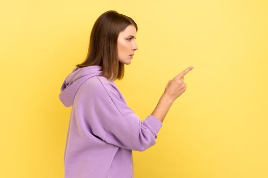 Hey you, be careful. side view portrait of young dark haired woman seriously pointing finger and looking ahead warning, wearing purple hoodie. Indoor studio shot isolated on yellow background.