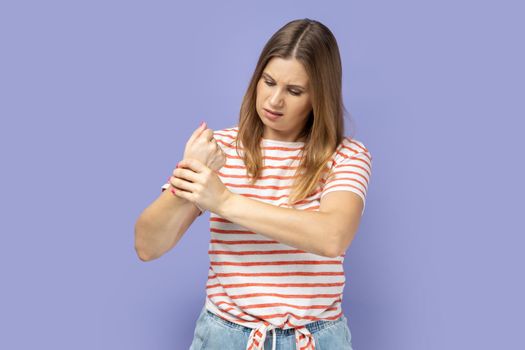 Unhappy sick blond woman wearing striped T-shirt touching painful hand, suffering trauma, sprain wrist, feeling ache of carpal tunnel syndrome. Indoor studio shot isolated on purple background.