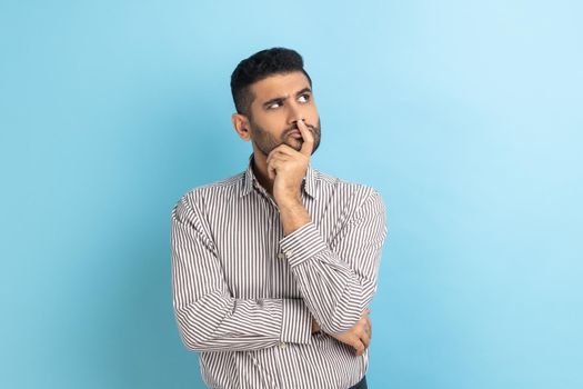 Portrait of thoughtful handsome bearded businessman holding his chin and pondering idea, confused not sure about solution, wearing striped shirt. Indoor studio shot isolated on blue background.